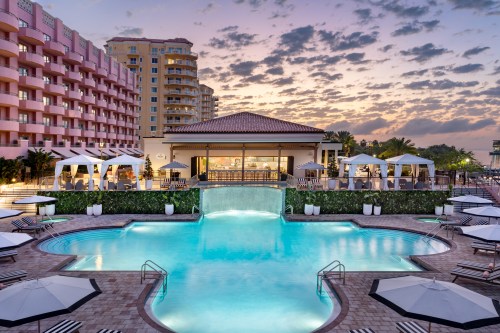A serene pool area with a waterfall, surrounded by lounge chairs and cabanas, set against a colorful sunset sky.
