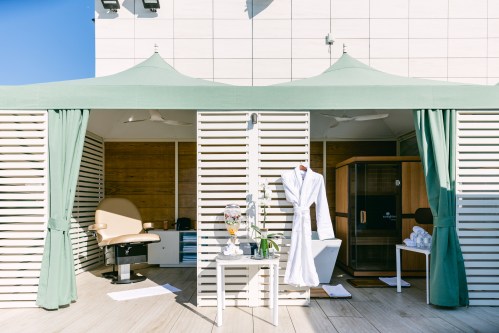 A spa-like outdoor area with green canopies, a chair, a robe, and a sauna, set against a clear blue sky.
