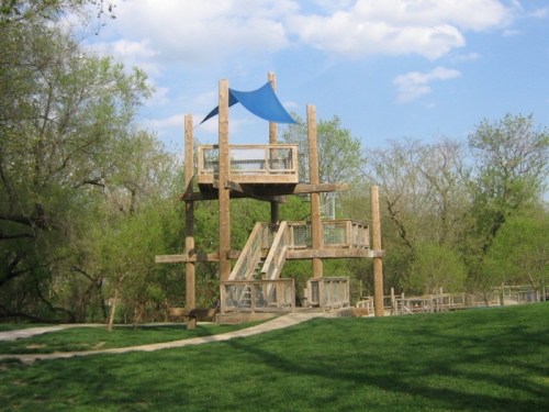 A wooden playground structure with a blue canopy, surrounded by green grass and trees under a clear blue sky.
