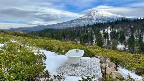 A vintage bathtub on a snowy terrace, surrounded by greenery and mountains under a cloudy sky.