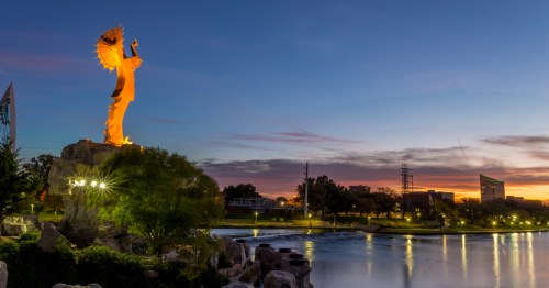 The Keeper of the Plains statue in Wichita, Kansas, is brightly lit as the sun rises over the city in the distance.