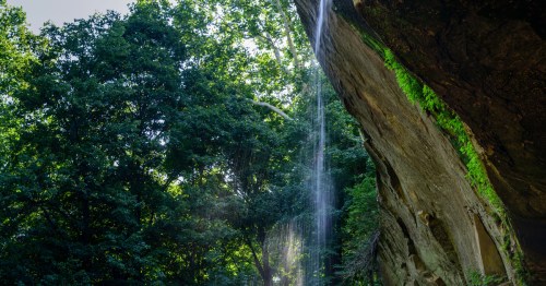 Sunlit waterfall spilling over a massive rock cliff in a lush green Indiana forest.