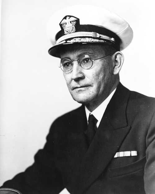 A man in a naval uniform and cap, wearing glasses, seated with a serious expression.