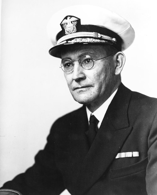 A man in a naval uniform and cap, wearing glasses, seated with a serious expression.