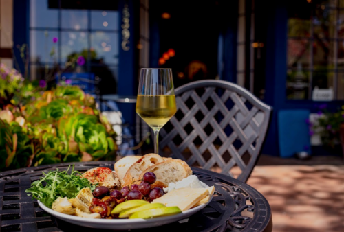 A plate of cheese, grapes, and bread beside a glass of white wine on a table outdoors, with a blue building in the background.