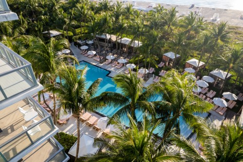 Aerial view of a tropical resort pool surrounded by palm trees and lounge chairs, near a sandy beach.
