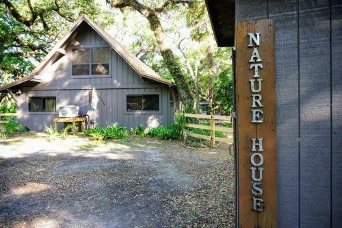 A wooden sign reading "Nature House" next to a cozy, gray cabin surrounded by trees and greenery.