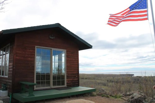 A wooden cabin with large windows, overlooking a landscape, with an American flag waving in the foreground.