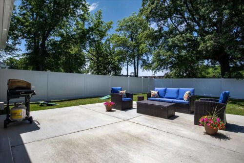 A sunny patio with blue outdoor furniture, a grill, and potted flowers, surrounded by green trees and a white fence.