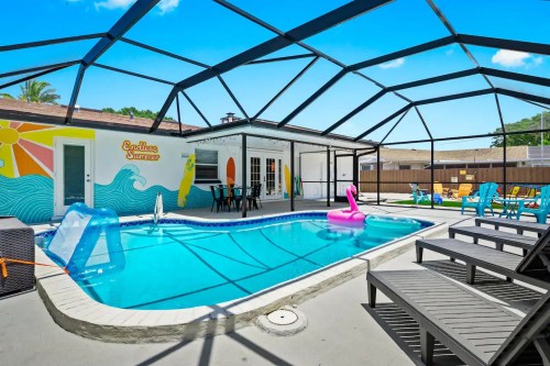 A bright pool area with a screened enclosure, colorful floats, and lounge chairs, featuring a mural of waves and surfboards.