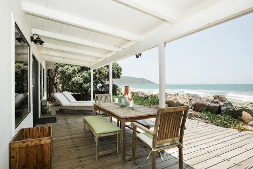 A coastal patio with a wooden table and chairs, overlooking the ocean and rocky shoreline under a white roof.