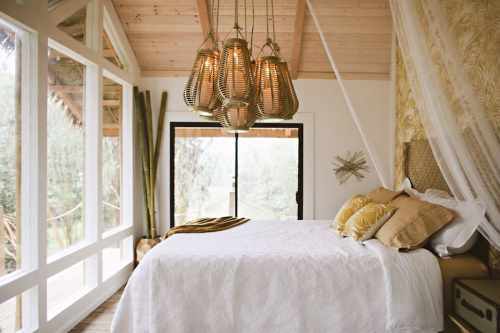 Cozy bedroom with a large bed, decorative pillows, and stylish pendant lights, featuring large windows and natural decor.