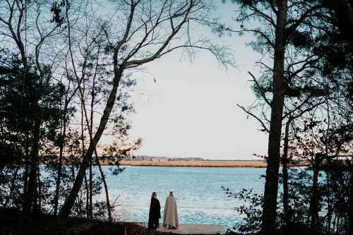 Two figures stand by a serene river, framed by trees, with a calm sky and distant landscape in the background.