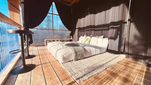 Cozy bedroom with a bed on a wooden floor, surrounded by sheer curtains and natural light.