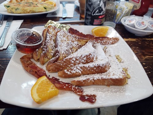 A plate of French toast dusted with powdered sugar, served with bacon, syrup, and an orange slice.