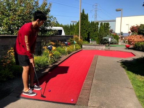 A person stands on a mini-golf course, preparing to putt on a red carpeted path surrounded by greenery and flowers.