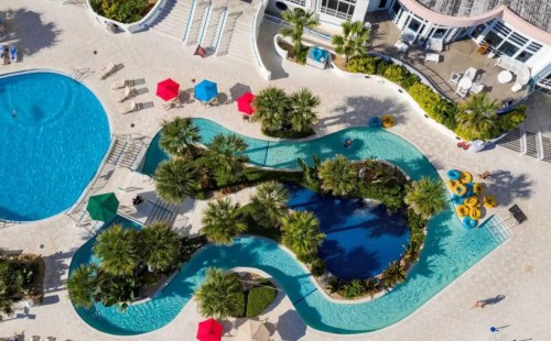 Aerial view of a resort pool area with winding water features, palm trees, and colorful umbrellas.