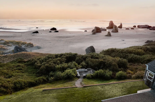 A serene beach scene at sunset, featuring rocks on the shore and lush greenery leading to a wooden pathway.
