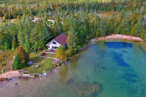 Aerial view of a lakeside house surrounded by trees and a sandy beach, with clear blue water and autumn foliage.