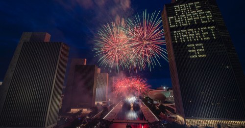 Colorful fireworks illuminate a city skyline at night, with buildings reflecting the vibrant display.