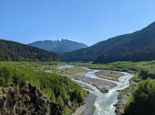 A winding river flows through a lush green valley, surrounded by mountains under a clear blue sky.