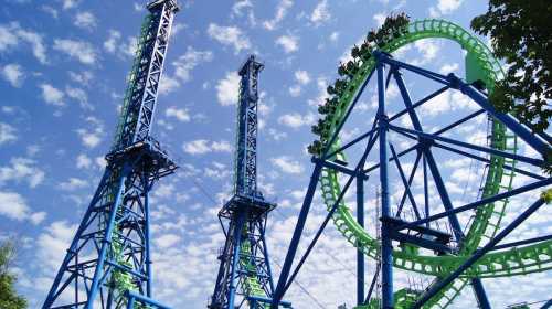 Two tall amusement park rides against a blue sky with scattered clouds, featuring a green and blue roller coaster.