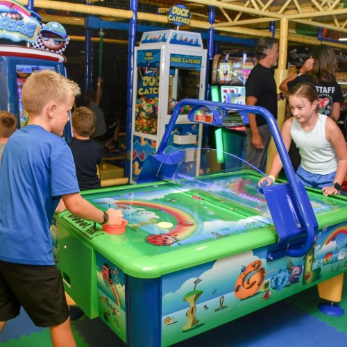 Two children play a colorful arcade game in a lively amusement center. Bright lights and other games are in the background.