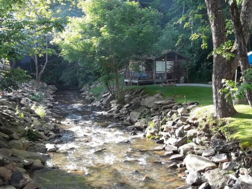 A serene stream flows past a cabin surrounded by trees and rocks, creating a peaceful natural setting.