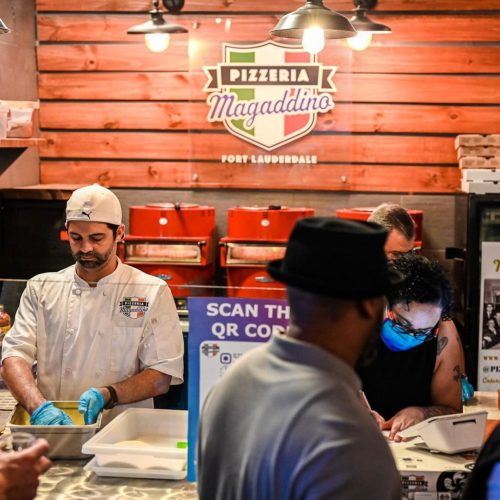 A chef prepares food in a bustling pizzeria, while customers engage at the counter, with a QR code sign visible.