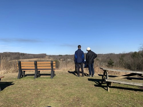 Two people stand on a grassy overlook, gazing at a scenic view with benches and picnic tables nearby.