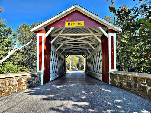 A red and white covered bridge with a stone entrance, surrounded by trees and a clear blue sky.