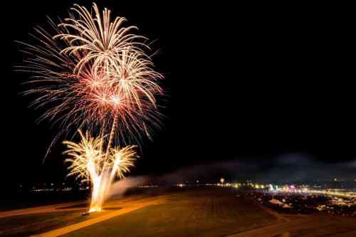 Colorful fireworks burst in the night sky over a festival, illuminating the dark landscape below.