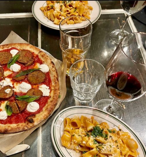 A table set with a pizza topped with meatballs and ricotta, pasta, glasses of beer, and red wine.