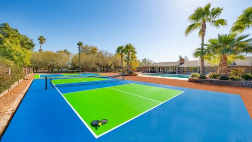 Bright outdoor tennis courts with blue and green surfaces, surrounded by palm trees and a pool area in the background.