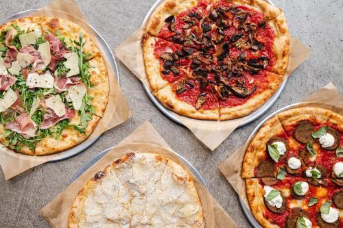 Four pizzas on metal trays: one with prosciutto and arugula, one with mushrooms, one with red sauce and toppings, and a dessert pizza.