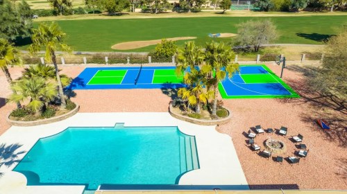 Aerial view of a pool, tennis court, and landscaped area with palm trees and a fire pit in a sunny outdoor setting.