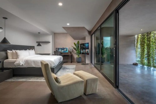 Modern bedroom with a large bed, a cozy chair, and glass doors leading to a patio with greenery.