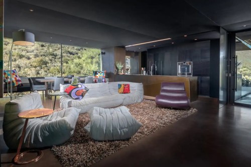 Modern living room with a plush sofa, colorful cushions, a sleek kitchen, and large windows overlooking greenery.
