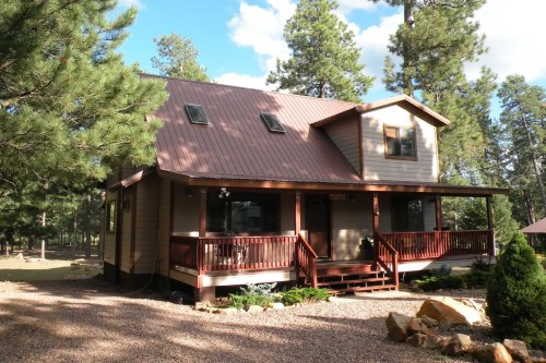 A two-story house with a brown metal roof, surrounded by trees and gravel, featuring a front porch and large windows.