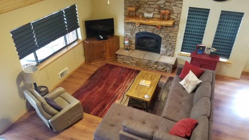 Cozy living room with a stone fireplace, wooden furniture, and large windows covered with dark blinds.