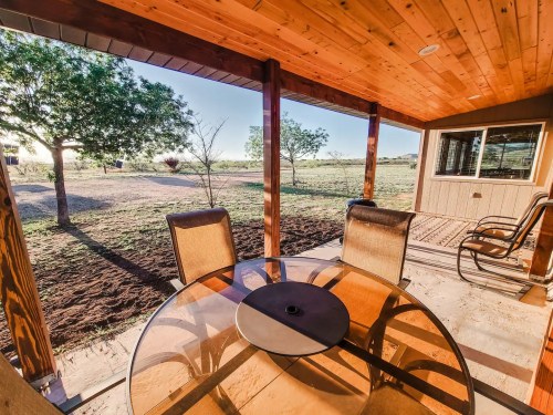 A patio with a glass table and chairs, overlooking a grassy area and trees in the background.