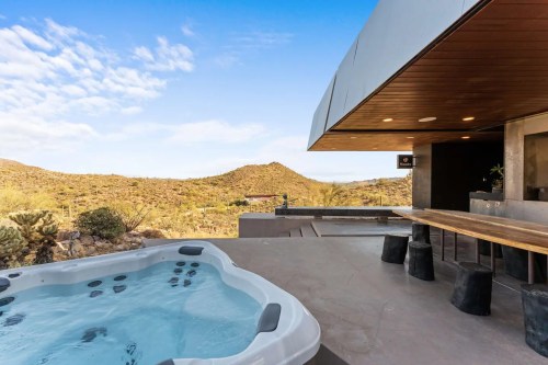 A modern outdoor space featuring a hot tub, wooden table, and scenic desert landscape under a blue sky.