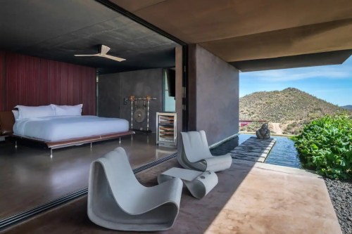 Modern bedroom with a large bed, sleek chairs, and a view of a desert landscape through open glass doors.