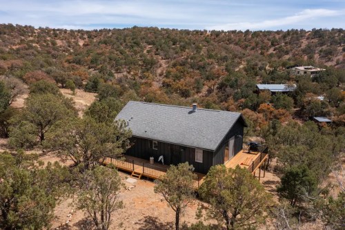 A secluded black cabin surrounded by trees on a hillside, with a wooden deck and a scenic landscape in the background.