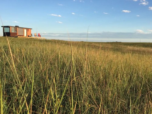 A modern cabin sits on a grassy hill under a blue sky, with distant fields stretching out in the background.