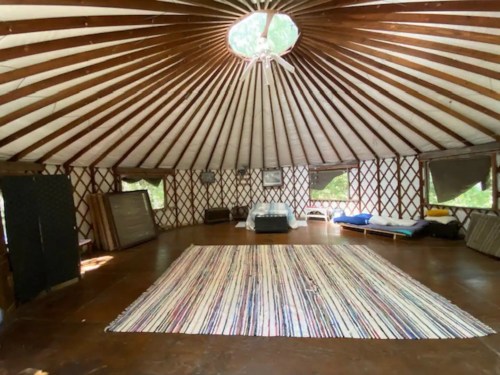 Interior of a yurt with a circular skylight, wooden beams, a large rug, and simple furnishings.