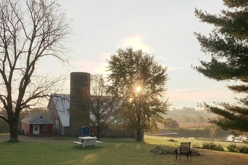 A serene farm scene at sunrise, featuring a barn, silo, trees, and a picnic table in a peaceful landscape.