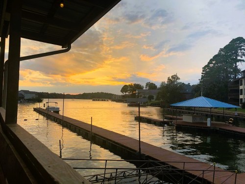 Sunset over a calm lake, with docks and boats, framed by trees and buildings under a colorful sky.