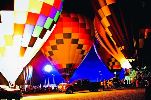 Colorful hot air balloons illuminated at night, with people and vehicles gathered below.