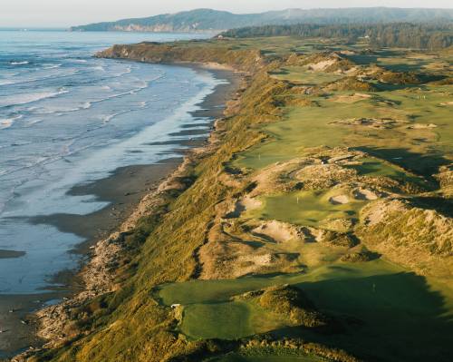 Aerial view of a coastal golf course with sandy areas, lush greens, and waves lapping at the shore in the background.